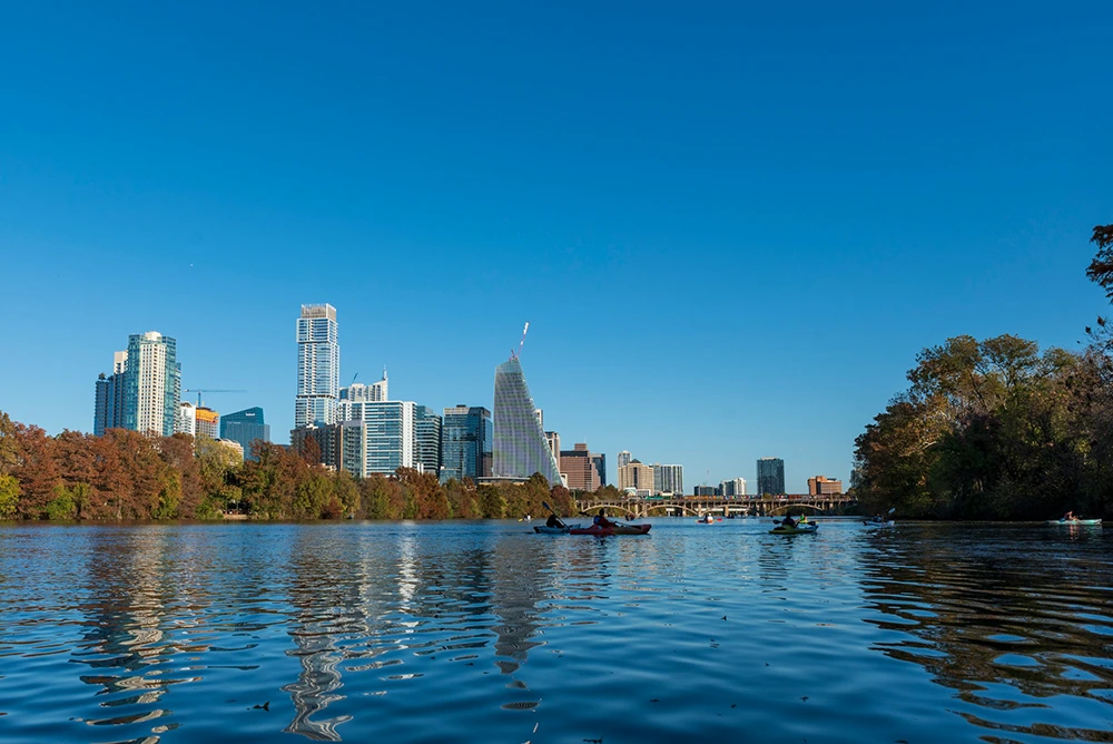 A serene lake foreground with people kayaking and the skyline of a modern city in the background under a clear blue sky.