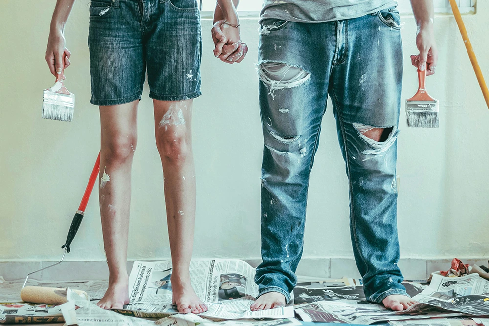 Two people in paint-splattered clothing holding paintbrushes with renovation supplies on the floor.