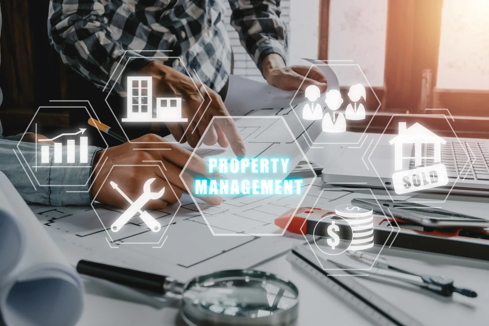 Alt text: "Concept image of property management with virtual graphics of houses, graphs, and maintenance tools over a real estate agent working at a desk with architectural plans and a laptop.